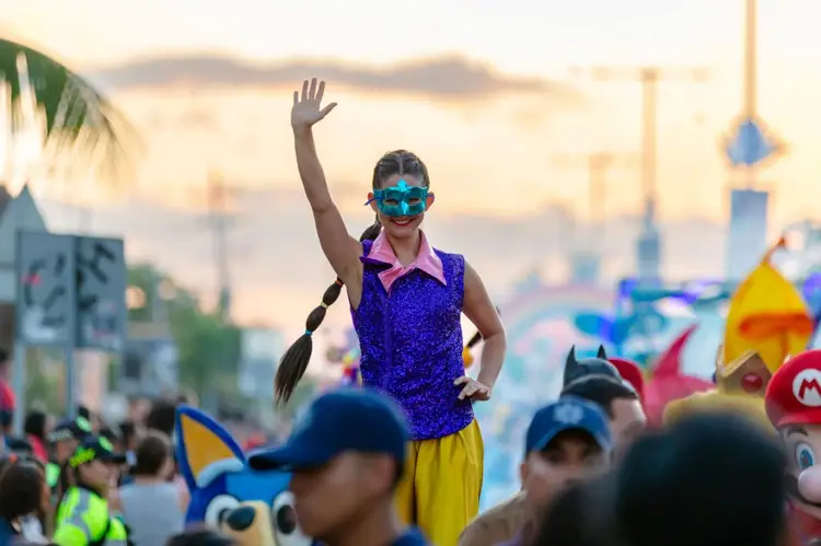 Person in blue sunglasses and purple sequined top waves from a float during a daytime parade with crowds and colorful character figures visible below