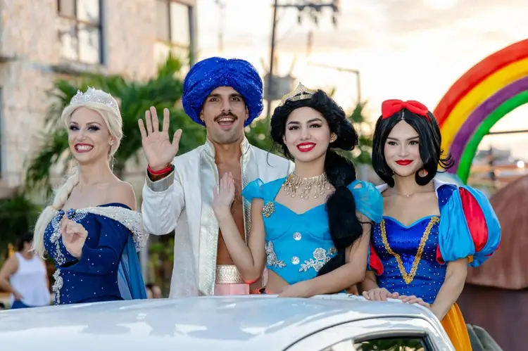 Four people in elaborate fairy tale costumes stand on a vehicle during a street celebration with a rainbow decoration visible behind them