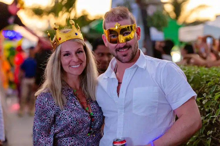Two people wearing gold masquerade masks pose together at an outdoor evening event with crowds and decorative lights in the background