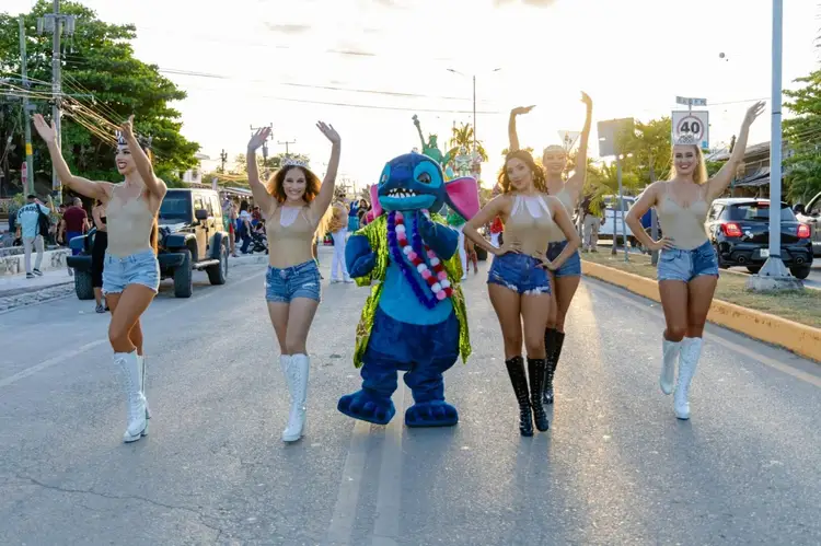 Performers in costume dance down a street during a daytime parade, with spectators lining the road and vehicles visible in the background