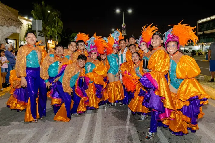 Group of children wearing colorful orange, blue and purple carnival costumes with large feathered headdresses posing together on a nighttime street