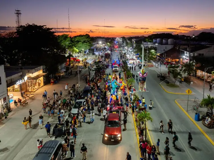 Crowds fill a street during an evening carnival celebration with decorated floats, vehicles, and illuminated vendor stalls under a twilight sky