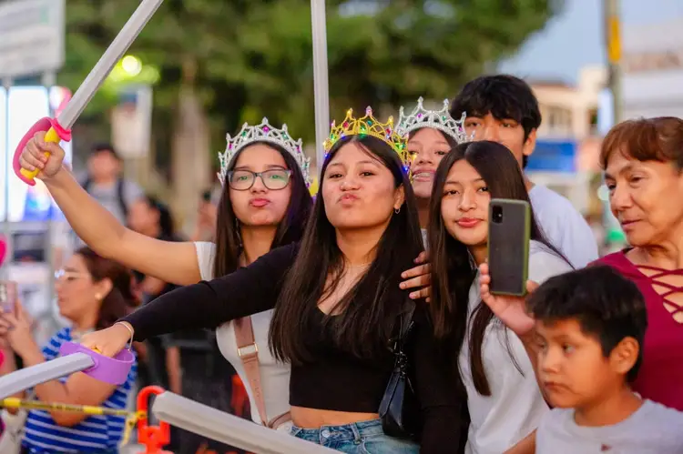 Group of young people wearing decorative crowns pose together at an outdoor event, with one holding up a selfie stick while others hold phones to record