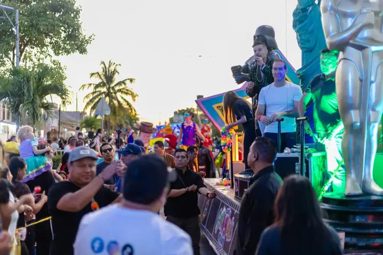 Performers on a stage with green lighting entertain a crowd gathered along a palm-lined street during a daytime carnival celebration