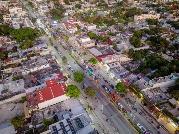 Aerial view of a crowded street festival with decorated floats and spectators lining both sides of a main avenue through a densely built urban neighborhood