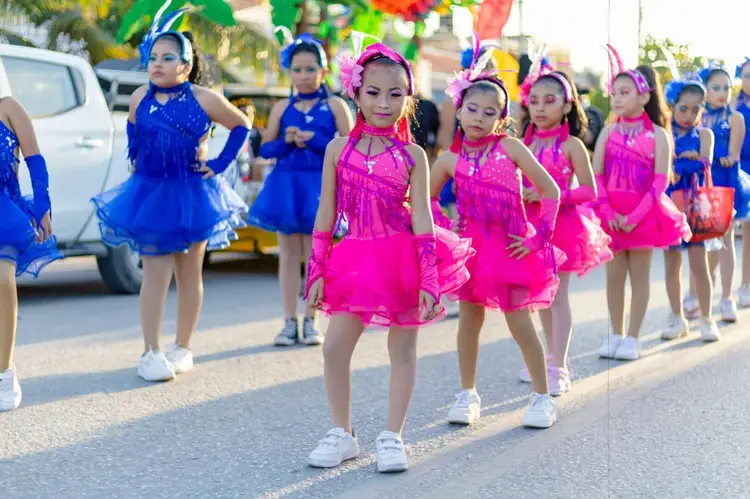 Children in blue and pink dance costumes with headbands perform in a parade on a street lined with parked vehicles