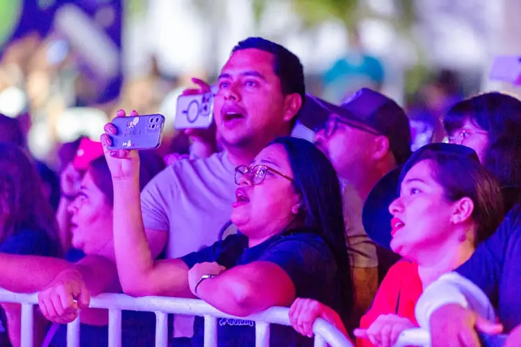 Crowd of people holding phones and looking up while standing behind a barrier at an outdoor event with purple and pink lighting