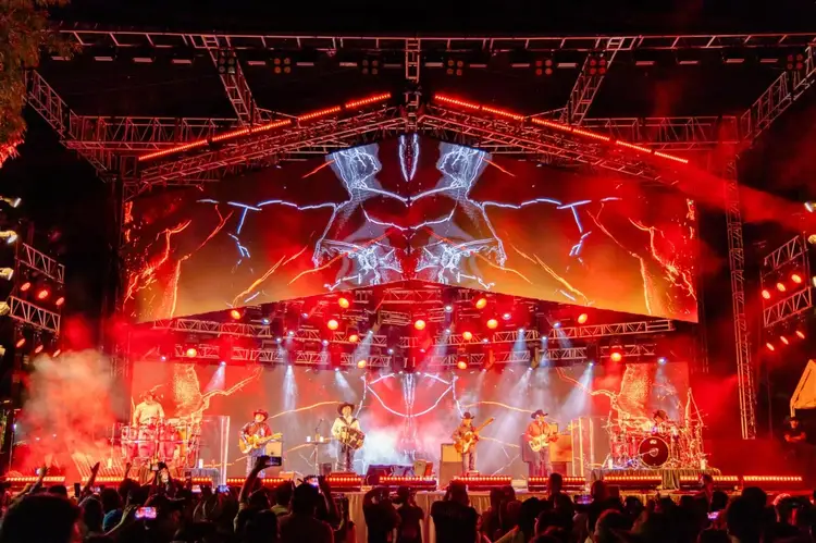 A band performs on stage under red lighting and lightning-effect visuals while a crowd watches with raised phones