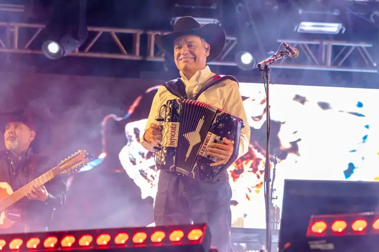 Musician in cowboy hat playing accordion on stage with guitarist and stage lighting in background