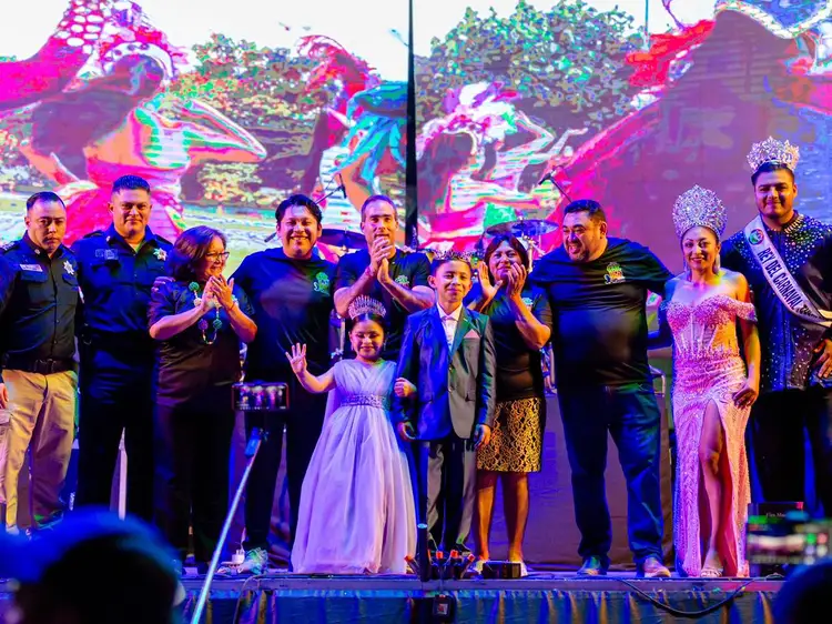 Group of officials, pageant contestants, and children stand together on stage under colorful lighting at what appears to be a carnival event