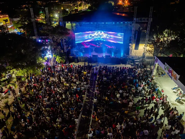 Aerial view of a large outdoor concert or festival venue at night with crowds gathered before a illuminated stage displaying visuals