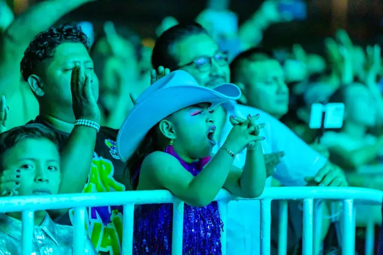 Spectators watch from a barrier at a nighttime event, with a child in a blue cowboy hat and purple outfit cheering in the foreground