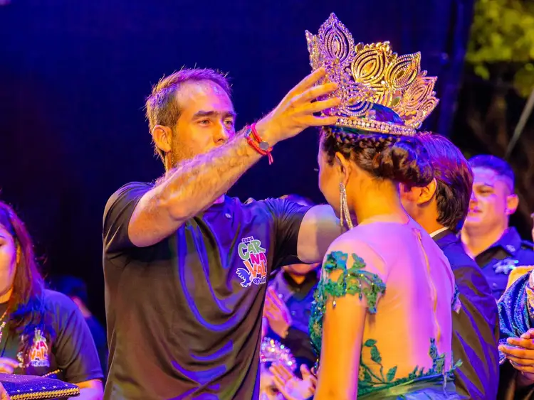 Man places an ornate jeweled crown on another person's head during a nighttime stage performance with onlookers around them