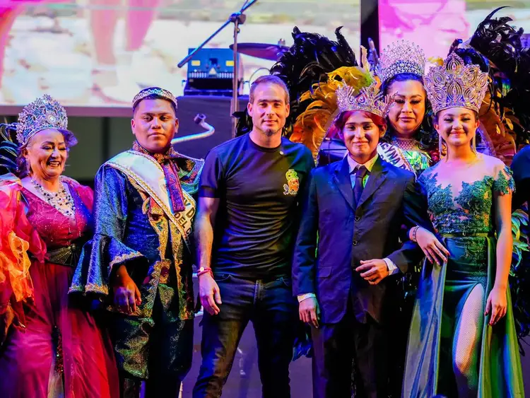 Group of performers in elaborate costumes with ornate headdresses and colorful clothing stand together on a stage under colored lighting
