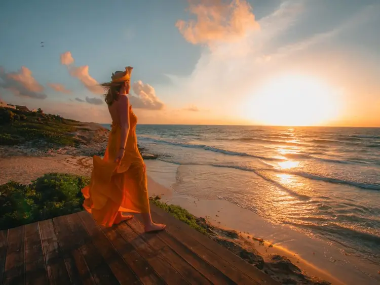 Woman in orange dress stands on wooden deck overlooking a beach at sunset with waves and cloudy sky