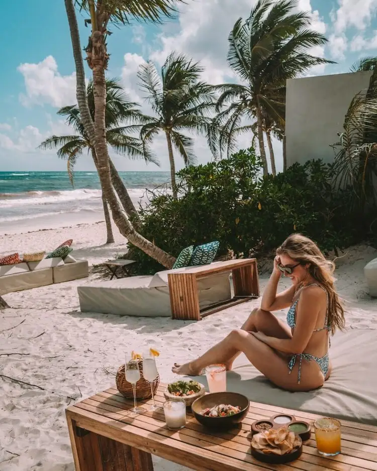 Woman in swimsuit sits on sand at a beachfront with food and drinks on a wooden table, palm trees and ocean visible behind her