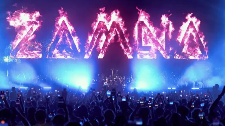 Crowd of concert attendees with raised hands and phones facing a stage lit by blue spotlights beneath large glowing letters spelling a word