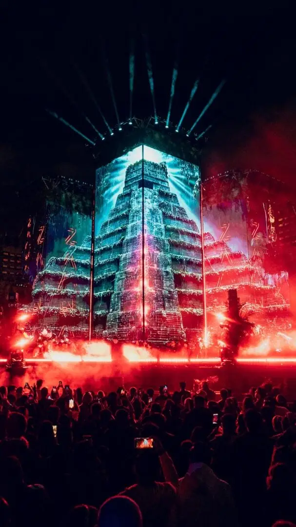 Crowd gathers beneath a tall building illuminated with blue and red neon lights during an outdoor nighttime event