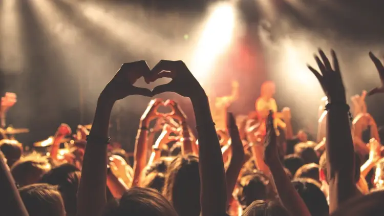 Concert attendee makes a heart shape with their hands while surrounded by a crowd with raised arms under stage lighting