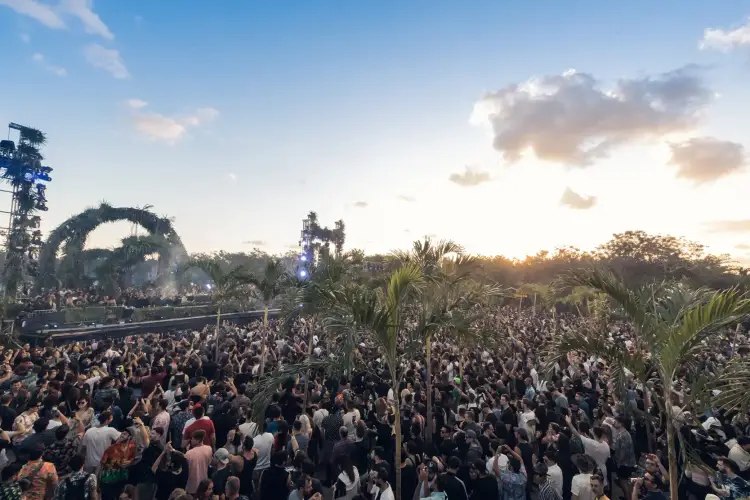Large outdoor music festival with crowd gathered around decorated stage structures and palm trees at dusk
