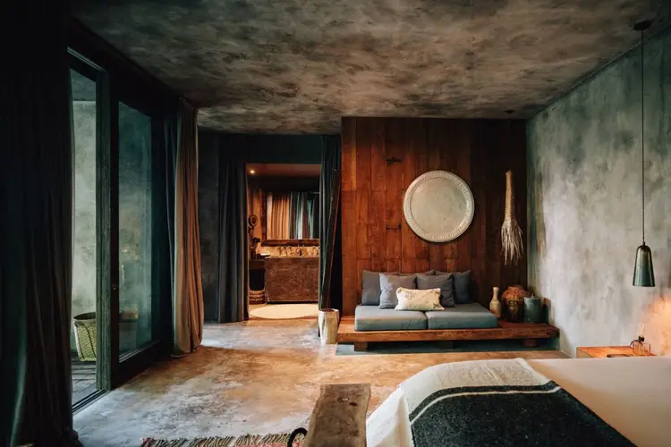 A modern hotel room with concrete walls and wooden accents features a low platform bed, sunken seating area, and large windows overlooking natural surroundings