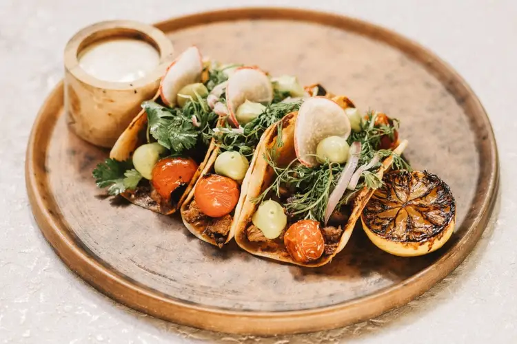Three tacos filled with roasted vegetables and greens sit on a wooden plate next to a small bowl of white sauce and charred lemon wedges