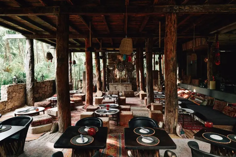 Open-air dining area with wooden beams and columns, black tables set with plates, woven pendant lights, and tropical vegetation visible through openings