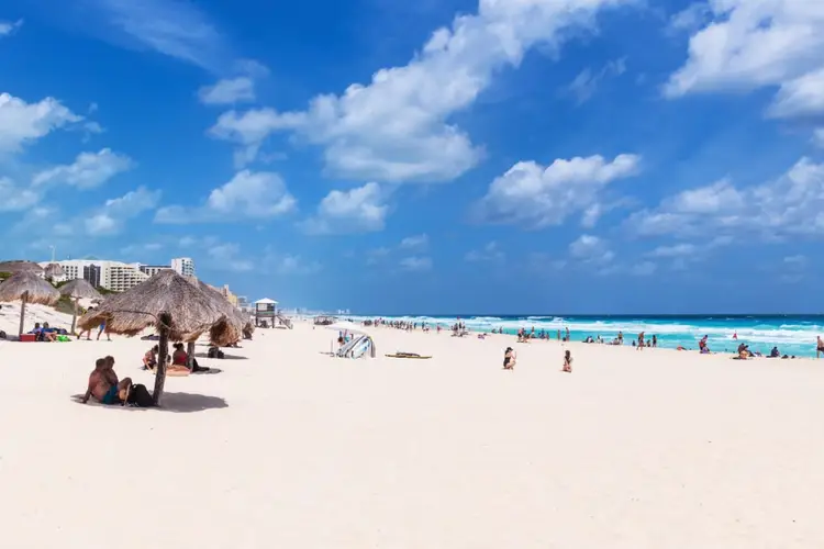 Beachgoers relax on sand under palapas and in turquoise water with high-rise hotels visible along the shoreline Beachgoers relax on sand under palapas and in turquoise water with high-rise hotels visible along the shoreline