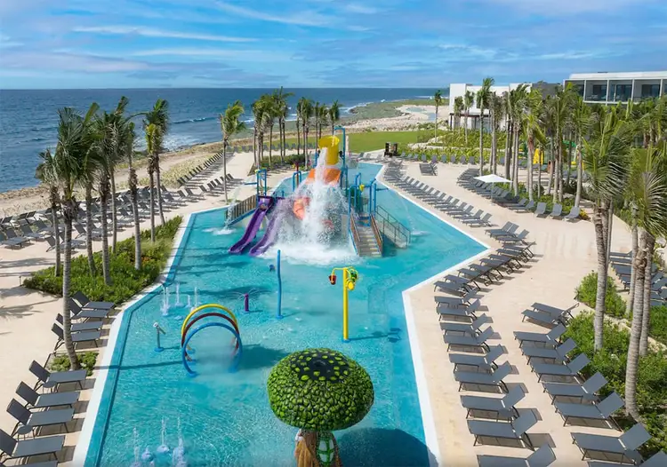 Aerial view of a resort waterpark with slides and shallow pools surrounded by lounge chairs, palm trees, and a beach with ocean beyond Aerial view of a resort waterpark with slides and shallow pools surrounded by lounge chairs, palm trees, and a beach with ocean beyond