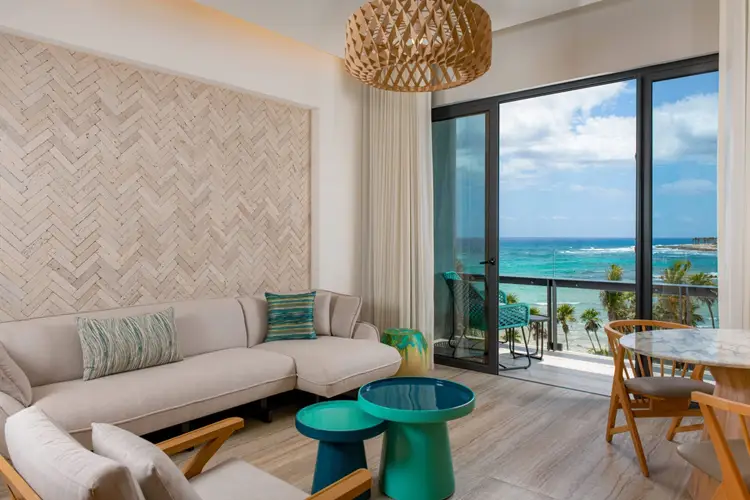 Modern hotel suite with beige sectional sofa, woven pendant light, and ocean view through floor-to-ceiling windows overlooking a tropical coastline Modern hotel suite with beige sectional sofa, woven pendant light, and ocean view through floor-to-ceiling windows overlooking a tropical coastline