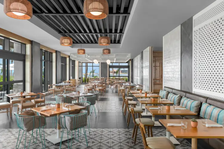 Modern restaurant interior with wooden tables, teal chairs, woven pendant lights, and floor-to-ceiling windows overlooking a coastal landscape Modern restaurant interior with wooden tables, teal chairs, woven pendant lights, and floor-to-ceiling windows overlooking a coastal landscape