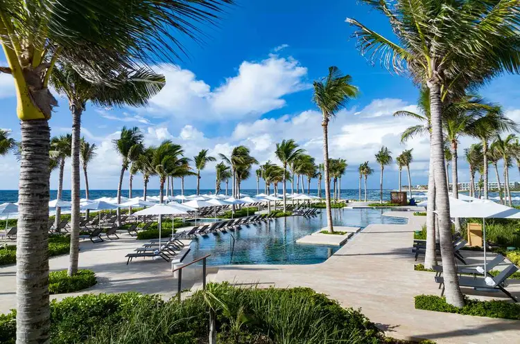 Resort pool lined with palm trees and lounge chairs, with ocean and white beach umbrellas visible in the background Resort pool lined with palm trees and lounge chairs, with ocean and white beach umbrellas visible in the background