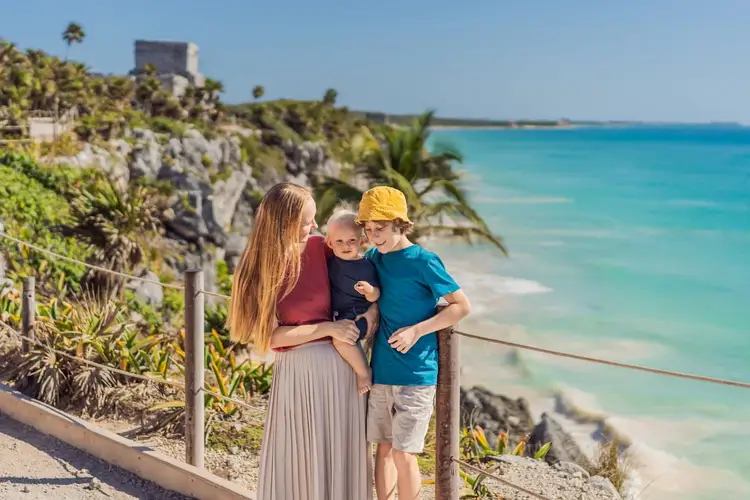 Woman and two children stand on a clifftop overlook above turquoise water and a beach, with tropical vegetation and a stone structure visible on the hillside be New Support Programs Boost Tourism in Tulum and Holbox