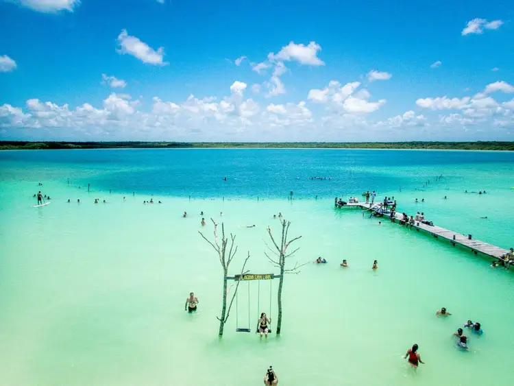 Swimmers and beachgoers wade and swim in turquoise shallow water near a wooden swing and a pier extending into the lagoon under blue sky with white clouds Swimmers and beachgoers wade and swim in turquoise shallow water near a wooden swing and a pier extending into the lagoon under blue sky with white clouds