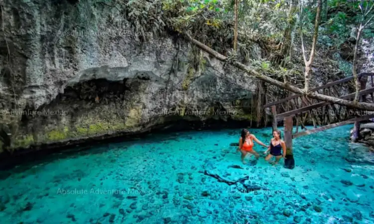 Swimmers wade in turquoise water beneath a cave opening in a jungle setting with wooden railings