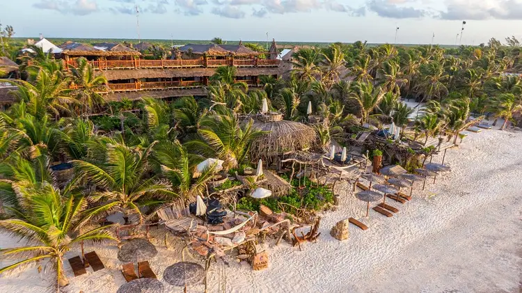 Beachfront resort with wooden structures, palm trees, and thatched-roof buildings overlooking sandy shore and water