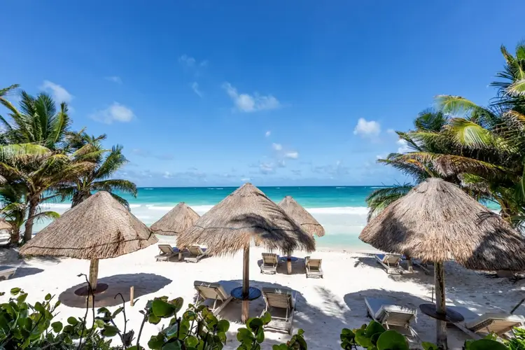 Beach resort with thatched-roof palapas, lounge chairs, and turquoise water under clear blue sky