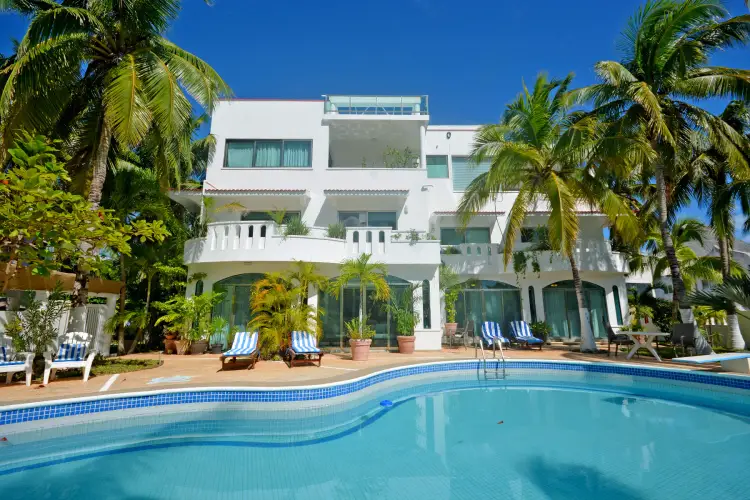 White multi-story residential building with swimming pool, lounge chairs, and palm trees under clear blue sky