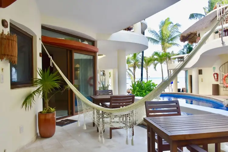 A hammock hangs on a beachfront property terrace with a wooden table, lounge chairs, and swimming pool visible nearby