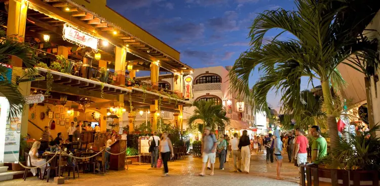 Pedestrians stroll through an open-air shopping and dining area lined with palm trees and illuminated colonial-style buildings at dusk