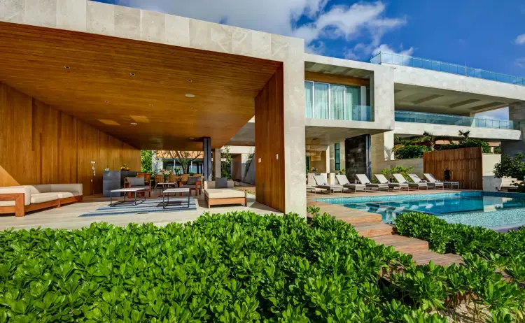 Modern house with wooden interior, lounge seating, and swimming pool surrounded by landscaping under blue sky