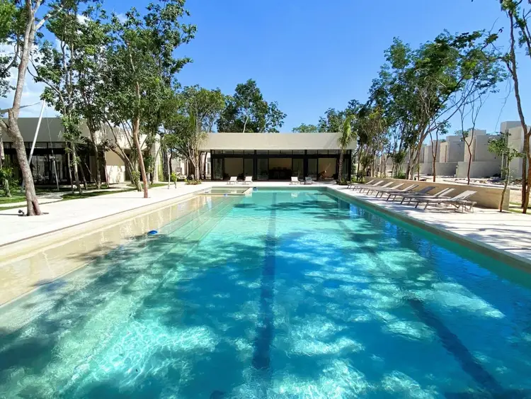 A rectangular swimming pool with clear turquoise water surrounded by loungers, mature trees, and modern beige buildings under a blue sky