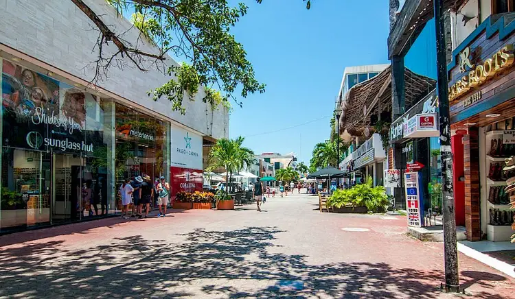 Pedestrians walk along a tree-lined outdoor shopping district with retail storefronts and brick paving on a clear day