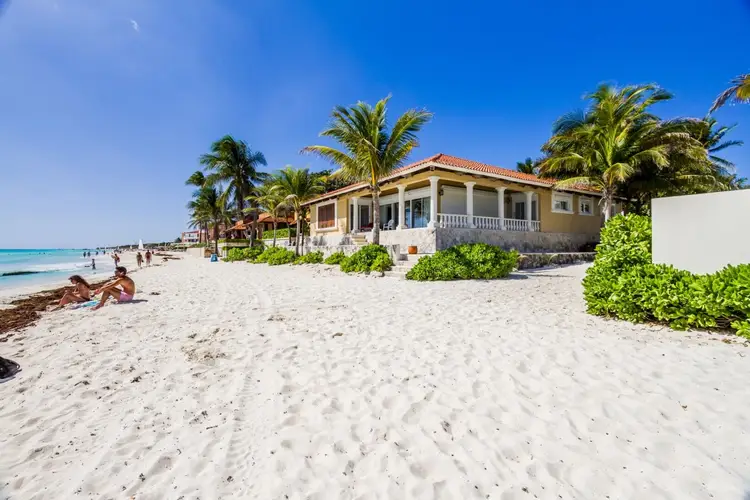 Yellow beachfront house with white columns and balustrade stands on sand beside turquoise water, with palm trees and beachgoers visible