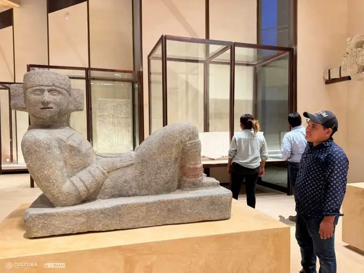 Visitors stand before a large stone pre-Columbian reclining figure sculpture displayed in a museum gallery with glass cases and beige walls Visitors stand before a large stone pre-Columbian reclining figure sculpture displayed in a museum gallery with glass cases and beige walls