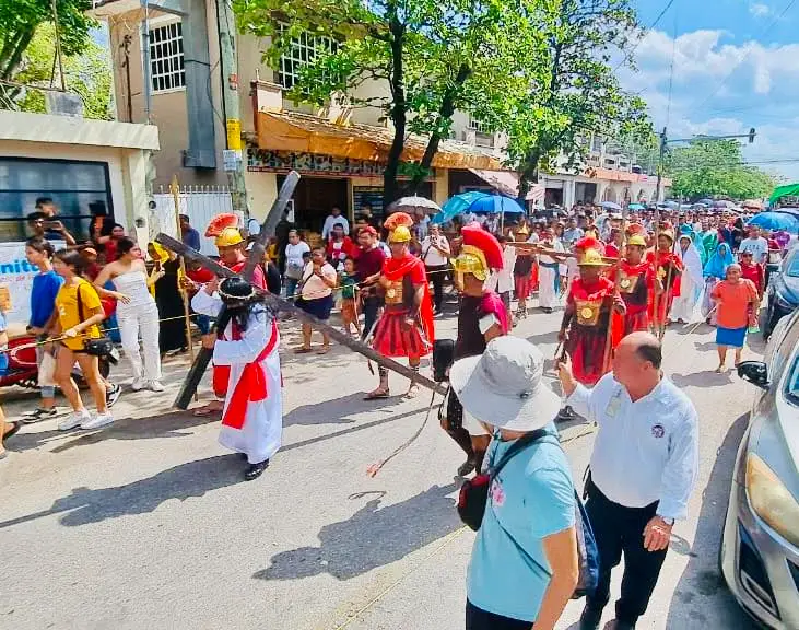 A large crowd gathers on a street for a religious procession, with participants wearing red and white robes and yellow hard hats walking among spectators A large crowd gathers on a street for a religious procession, with participants wearing red and white robes and yellow hard hats walking among spectators