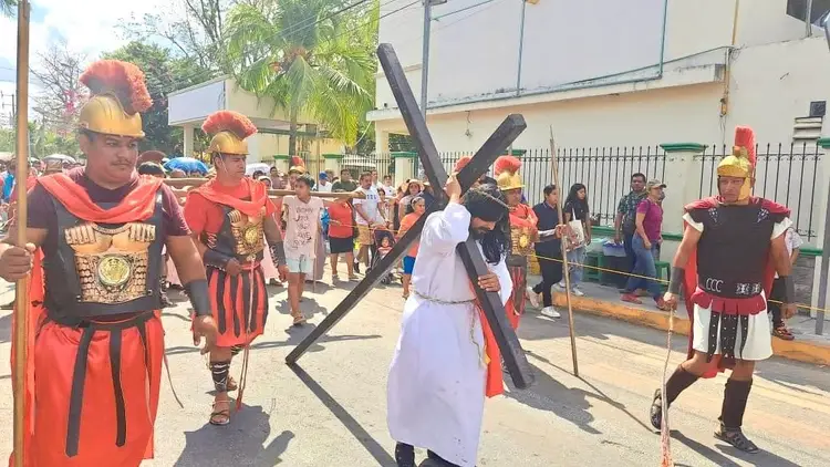 Person in white robe carrying large wooden cross walks down street surrounded by participants in Roman soldier costumes during a religious procession Person in white robe carrying large wooden cross walks down street surrounded by participants in Roman soldier costumes during a religious procession