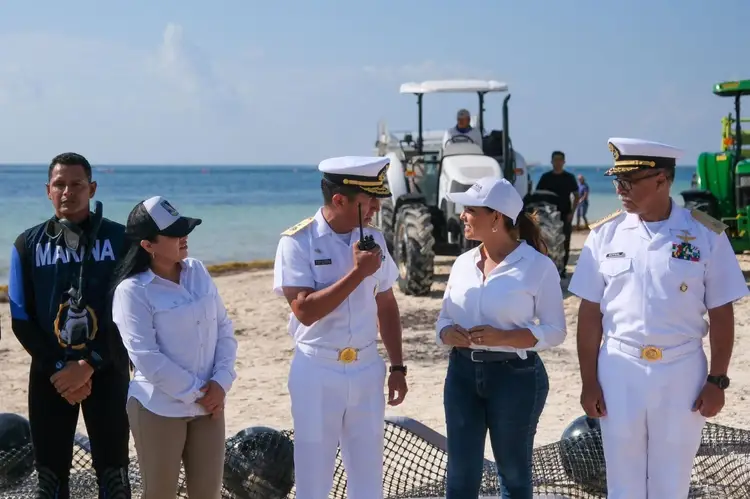 Military and civilian personnel stand on a beach near fishing nets and a tractor, with ocean and additional workers visible in the background