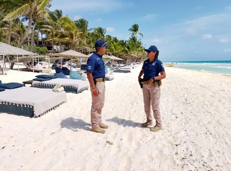Two uniformed officials stand on a beach near rows of lounge chairs and thatched umbrellas, with palm trees and turquoise water visible in the background Guard Deployed to Secure Quintana Roo Beaches During Easter Holiday