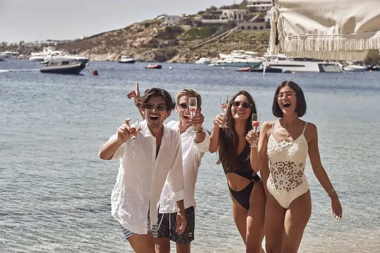 Four people in swimwear and light clothing stand in shallow water holding glasses, with moored boats and hillside buildings visible behind them